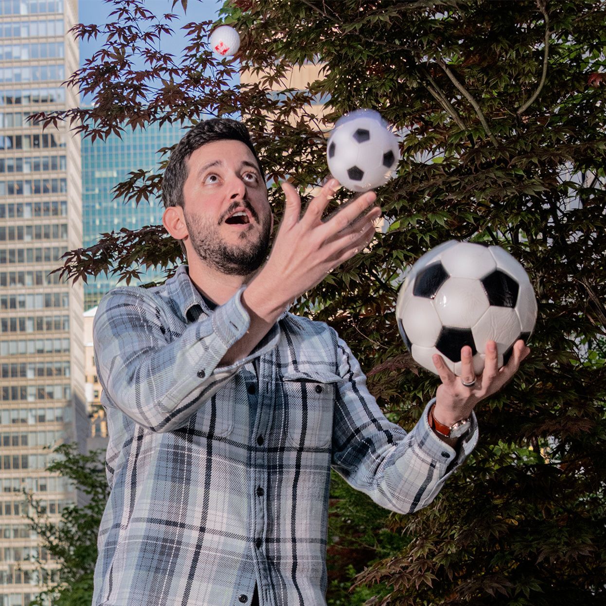 Man juggling soccer balls outdoors in front of trees and city buildings