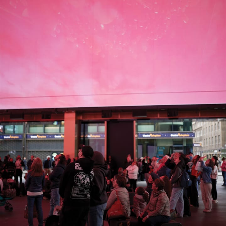 People stand staring up at a pink digital screen outside.