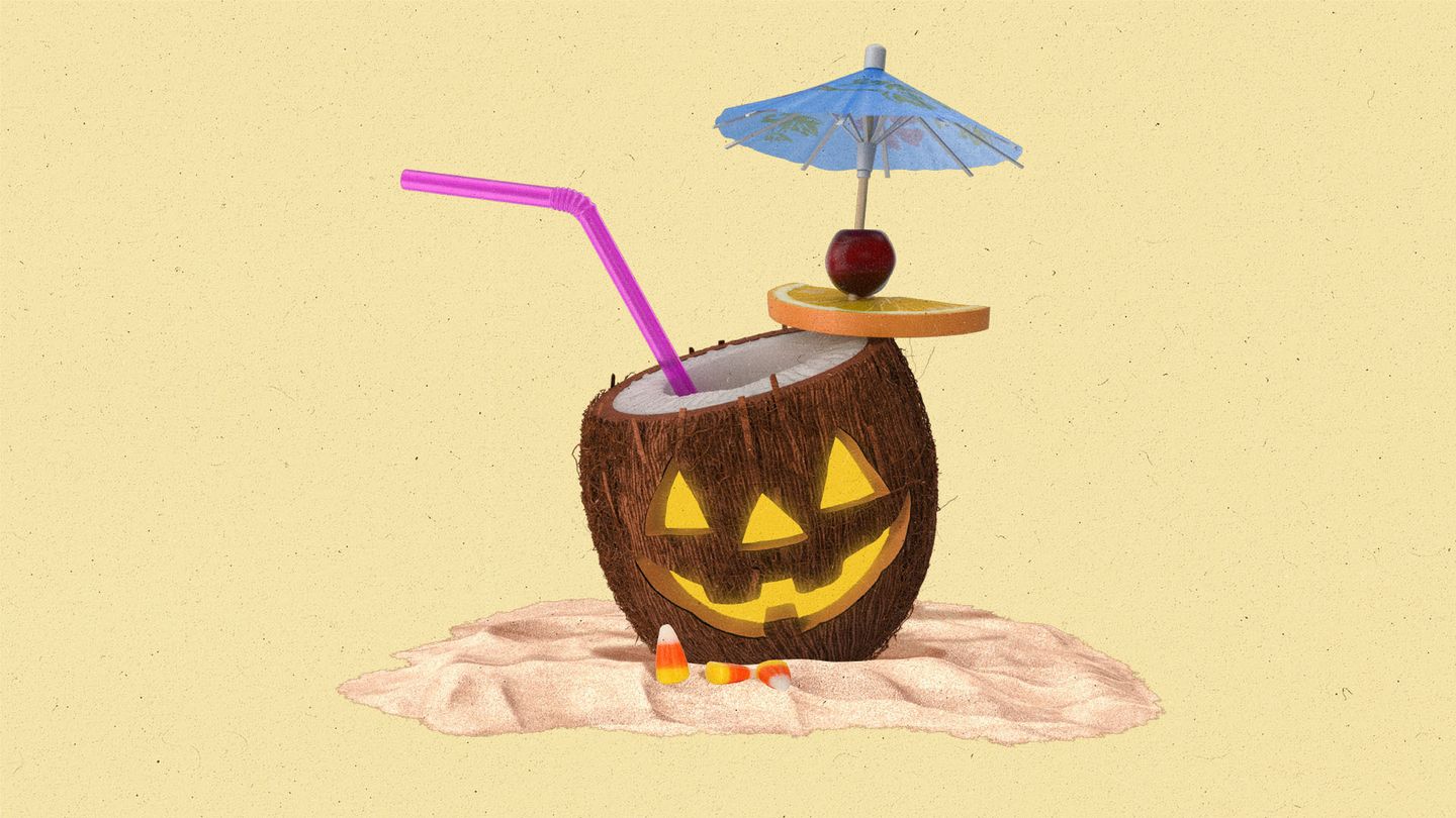 Open coconut sitting in the sand with a straw and mini umbrella with a jack-o-lantern face carved into it.
