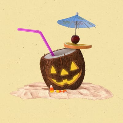 Open coconut sitting in the sand with a straw and mini umbrella with a jack-o-lantern face carved into it.