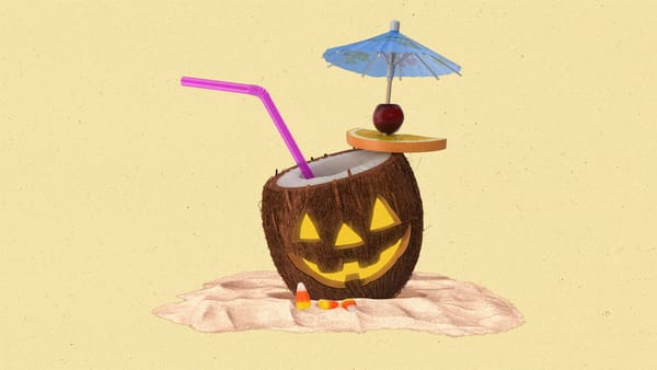 Open coconut sitting in the sand with a straw and mini umbrella with a jack-o-lantern face carved into it.